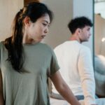 A man and woman sitting on a bed in a bedroom, looking away, suggesting tension.