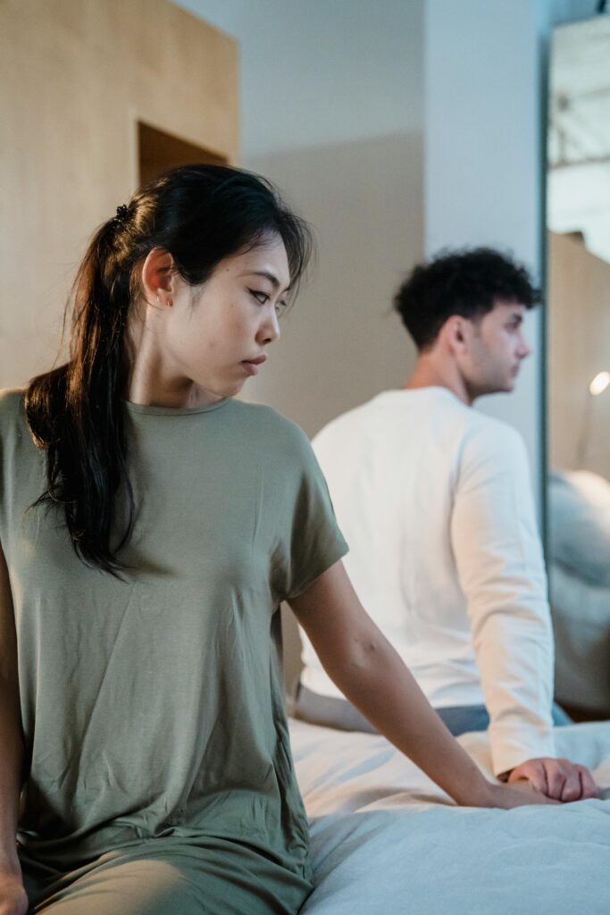 A man and woman sitting on a bed in a bedroom, looking away, suggesting tension.